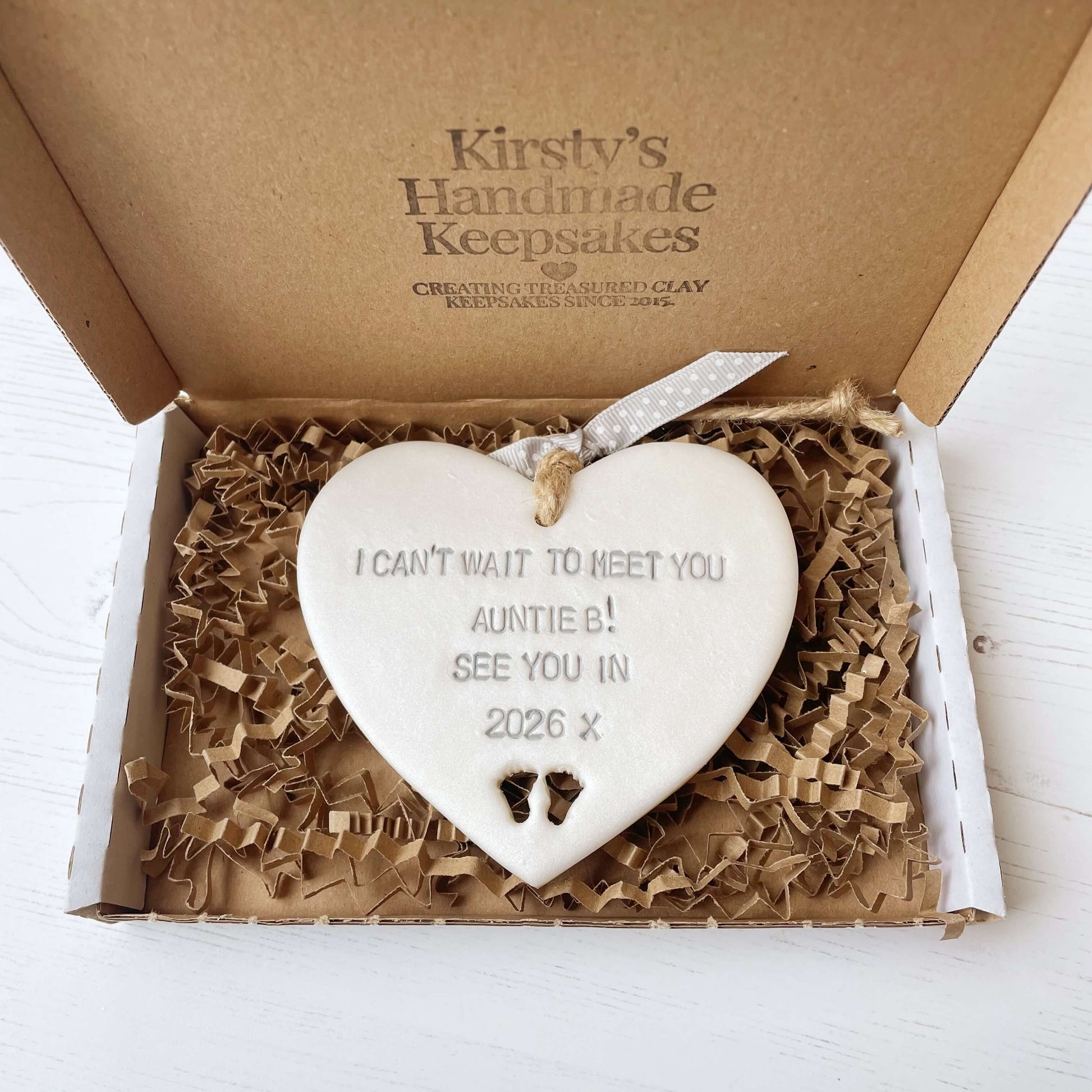 Pearlised white clay hanging heart with baby feet cut out of the bottom and grey personalisation, the heart is personalised with I CAN’T WAIT TO MEET YOU AUNTIE B! SEE YOU IN 2026 X