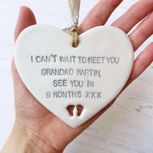 Pearlised white clay hanging heart with baby feet cut out of the bottom and grey personalisation, the heart is personalised with I CAN’T WAIT TO MEET YOU GRANDAD MARTIN, SEE YOU IN 9 MONTHS XXX