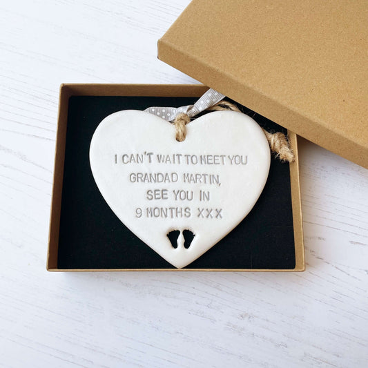 Pearlised white clay hanging heart with baby feet cut out of the bottom and grey personalisation, the heart is personalised with I CAN’T WAIT TO MEET YOU GRANDAD MARTIN, SEE YOU IN 9 MONTHS XXX