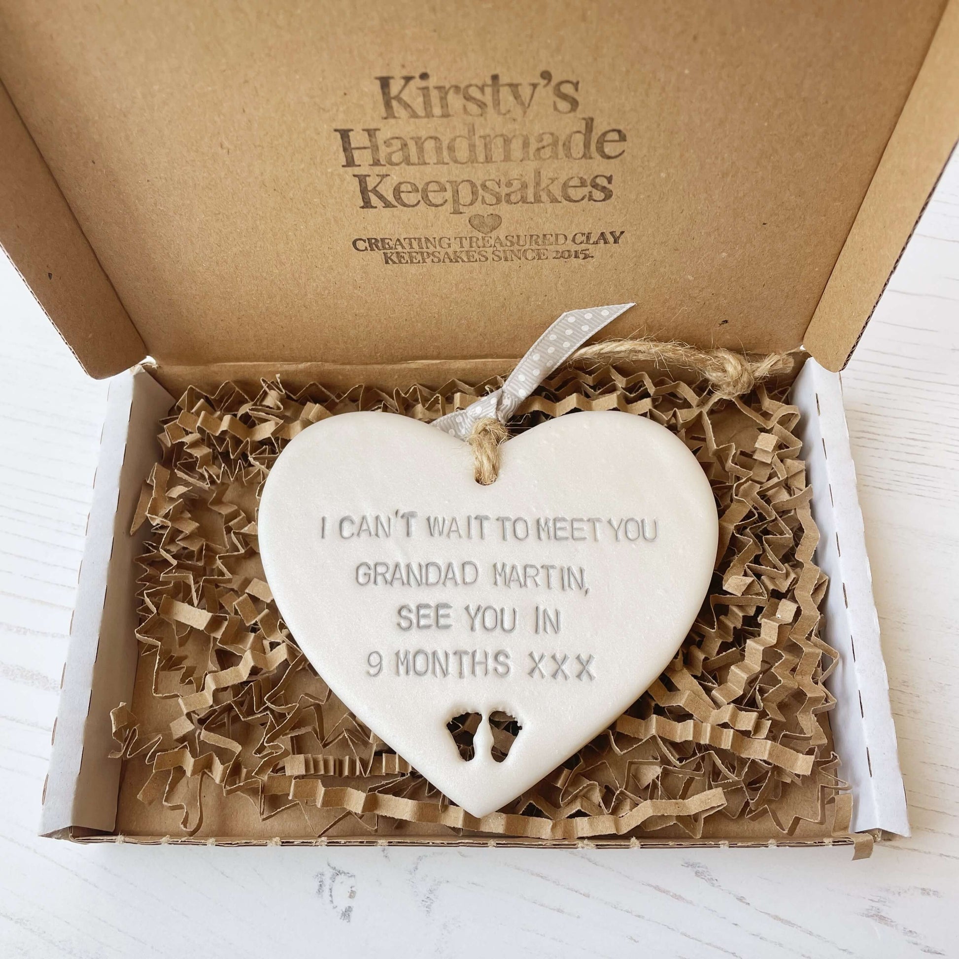 Pearlised white clay hanging heart with baby feet cut out of the bottom and grey personalisation, the heart is personalised with I CAN’T WAIT TO MEET YOU GRANDAD MARTIN, SEE YOU IN 9 MONTHS XXX