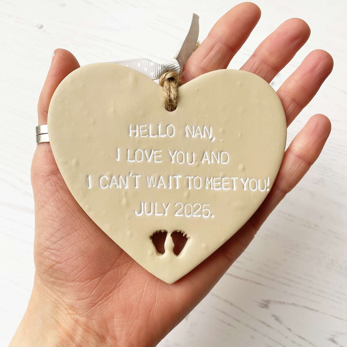 Beige clay hanging heart with baby feet cut out of the bottom and white personalisation, the heart is personalised with HELLO NAN, I LOVE YOU AND I CAN’T WAIT TO MEET YOU! JULY 2025.