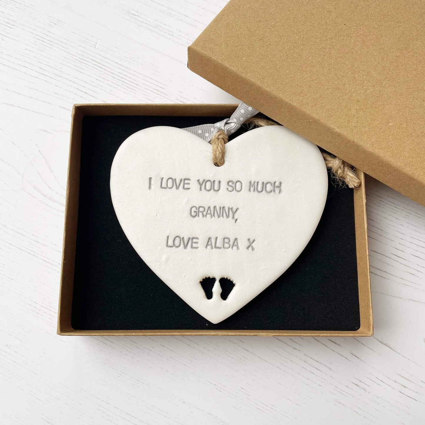 Pearlised white clay hanging heart with baby feet cut out of the bottom and grey personalisation, the heart is personalised with I LOVE YOU SO MUCH GRANNY, LOVE ALBA X