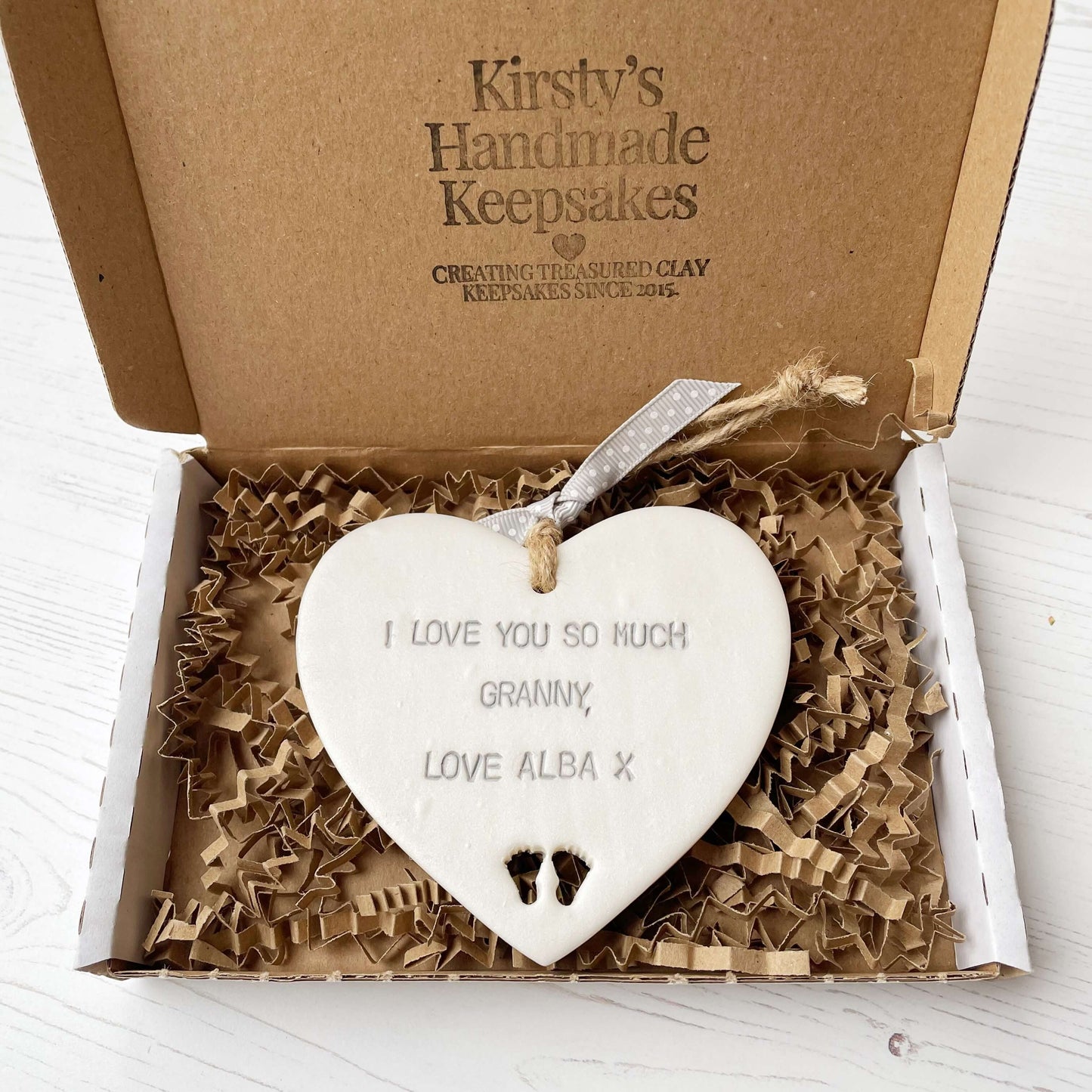 Pearlised white clay hanging heart with baby feet cut out of the bottom and grey personalisation, the heart is personalised with I LOVE YOU SO MUCH GRANNY, LOVE ALBA X