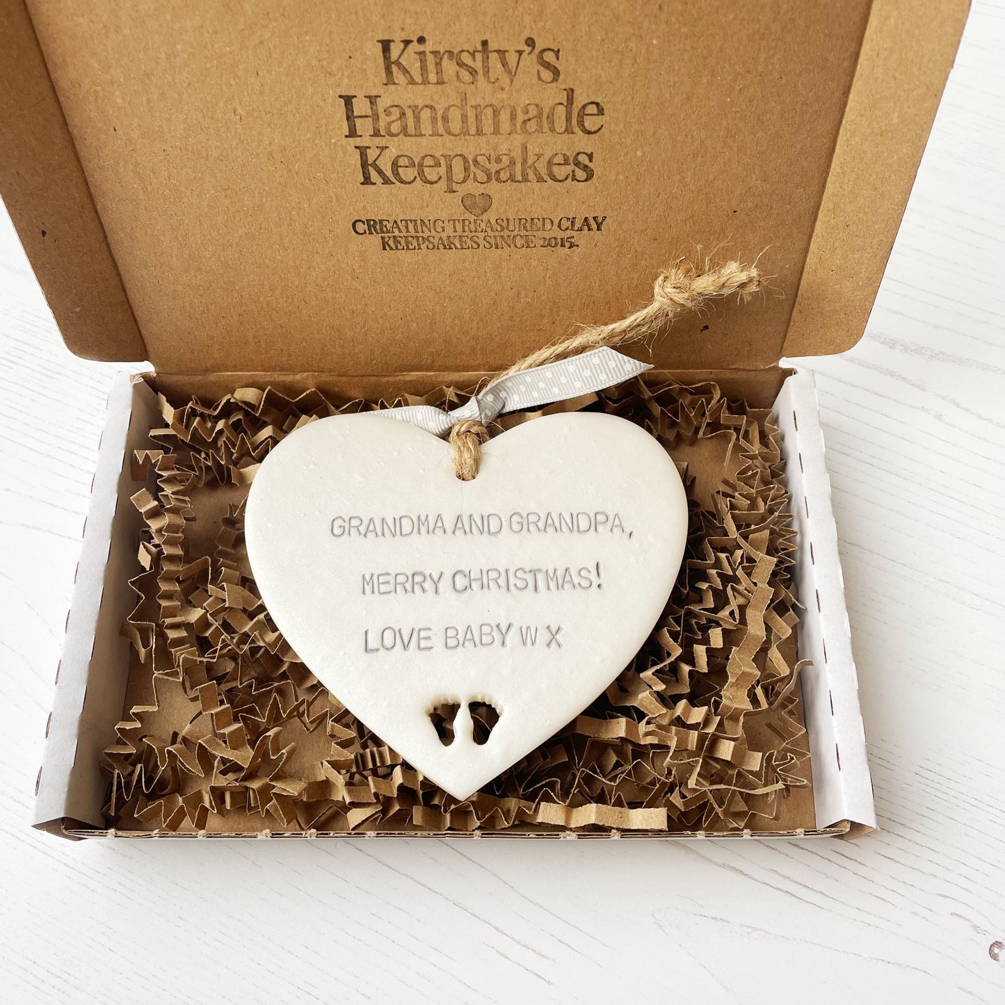 Pearlised white clay hanging heart with baby feet cut out of the bottom and grey personalisation, the heart is personalised with GRANDMA & GRANDPA, MERRY CHRISTMAS! LOVE BABY W X