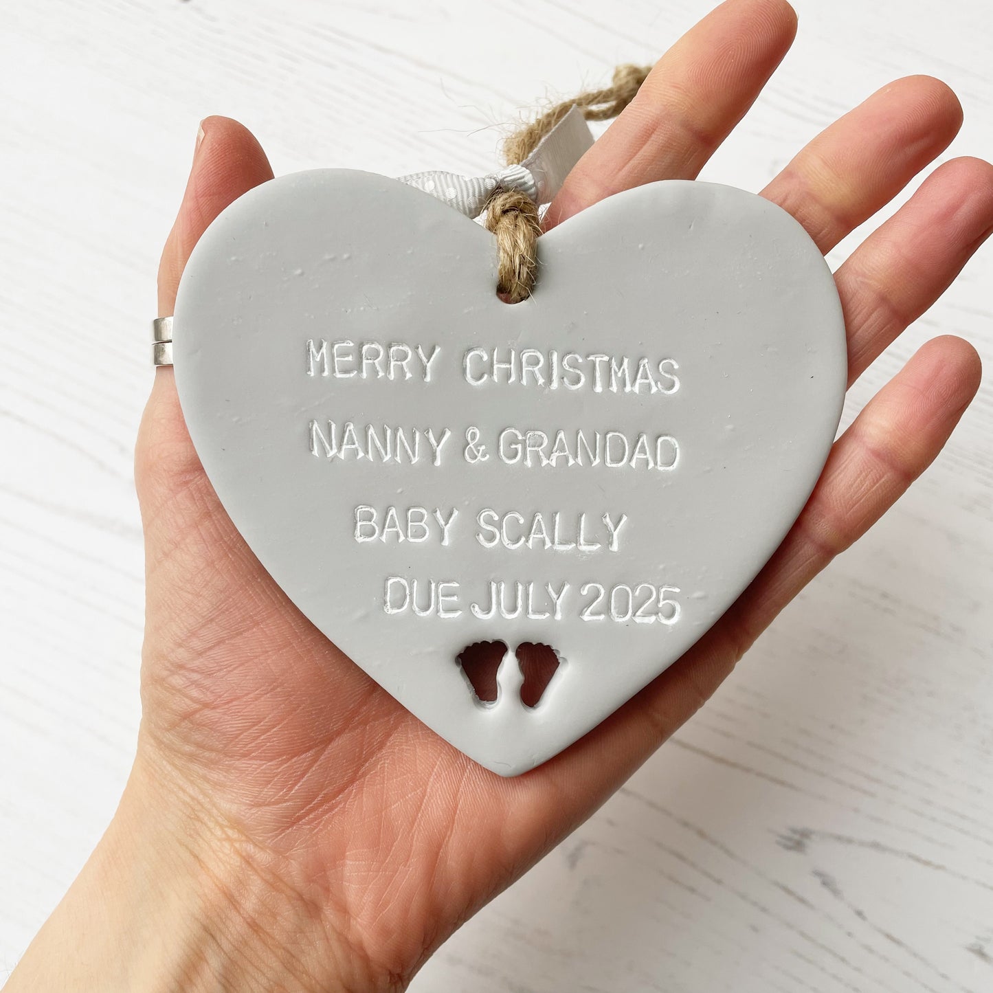Grey clay hanging heart with baby feet cut out of the bottom and white personalisation, the heart is personalised with MERRY CHRISTMAS NANNY & GRANDAD BABY SCALLY DUE JULY 2025