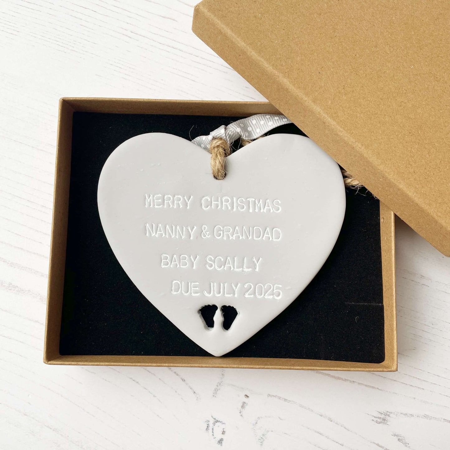Grey clay hanging heart with baby feet cut out of the bottom and white personalisation, the heart is personalised with MERRY CHRISTMAS NANNY & GRANDAD BABY SCALLY DUE JULY 2025