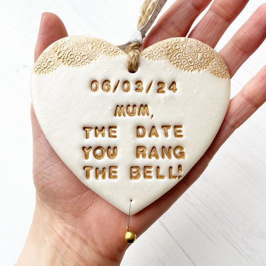 Personalised cancer survivor gift, pearlised white clay hanging heart with a gold lace edge at the top of the heart and a gold bell hanging below, the heart is personalised with 06/03/24 MUM, THE DATE YOU RANG THE BELL!