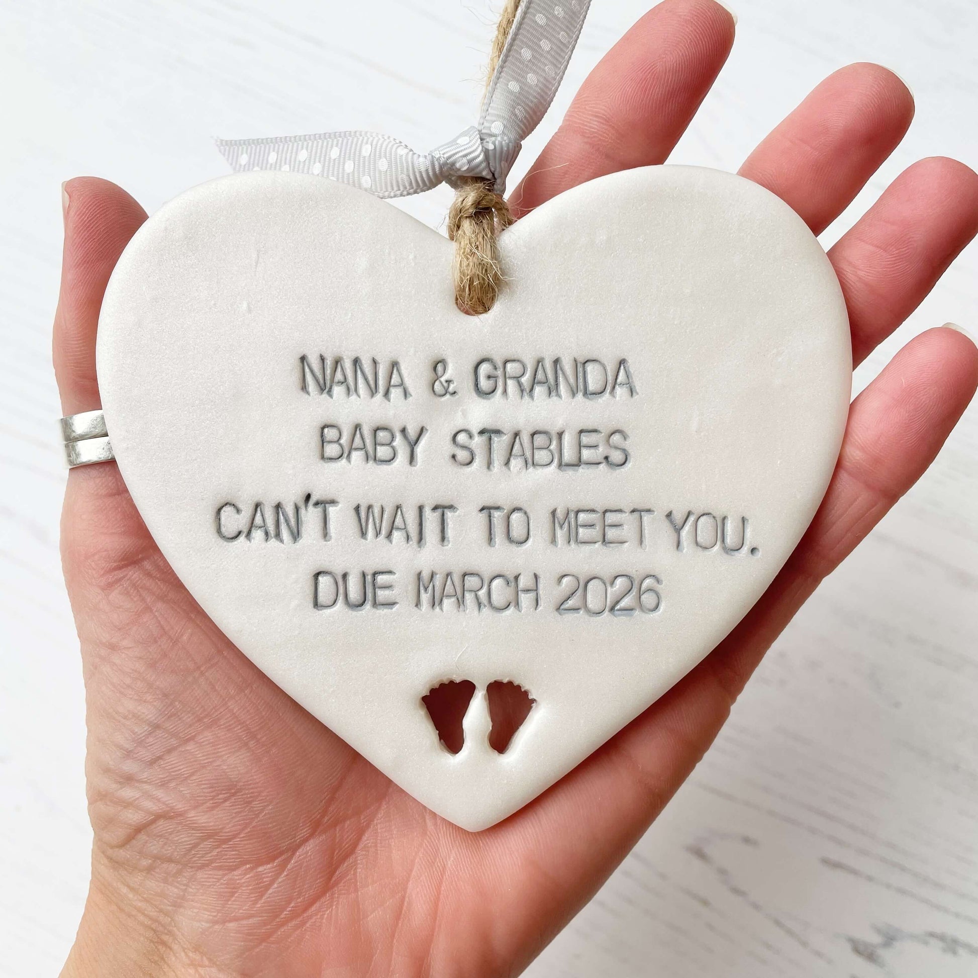 Pearlised white clay hanging heart with baby feet cut out of the bottom and grey personalisation, the heart is personalised with NANA & GRANDA BABY STABLES CAN’T WAIT TO MEET YOU DUE MARCH 2026