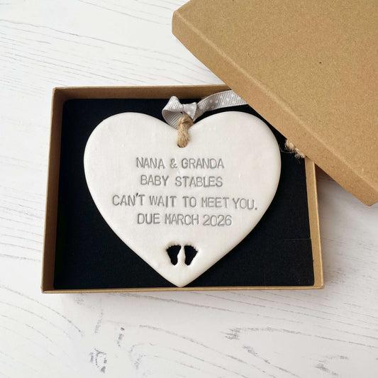 Pearlised white clay hanging heart with baby feet cut out of the bottom and grey personalisation, the heart is personalised with NANA & GRANDA BABY STABLES CAN’T WAIT TO MEET YOU DUE MARCH 2026