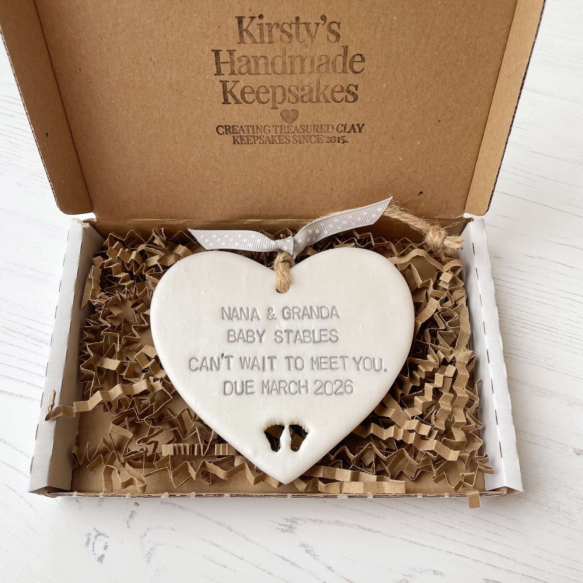 Pearlised white clay hanging heart with baby feet cut out of the bottom and grey personalisation, the heart is personalised with NANA & GRANDA BABY STABLES CAN’T WAIT TO MEET YOU DUE MARCH 2026