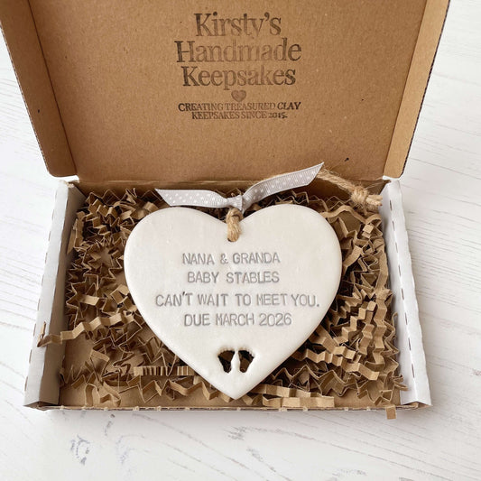 Pearlised white clay hanging heart with baby feet cut out of the bottom and grey personalisation, the heart is personalised with NANA & GRANDA BABY STABLES CAN’T WAIT TO MEET YOU DUE MARCH 2026