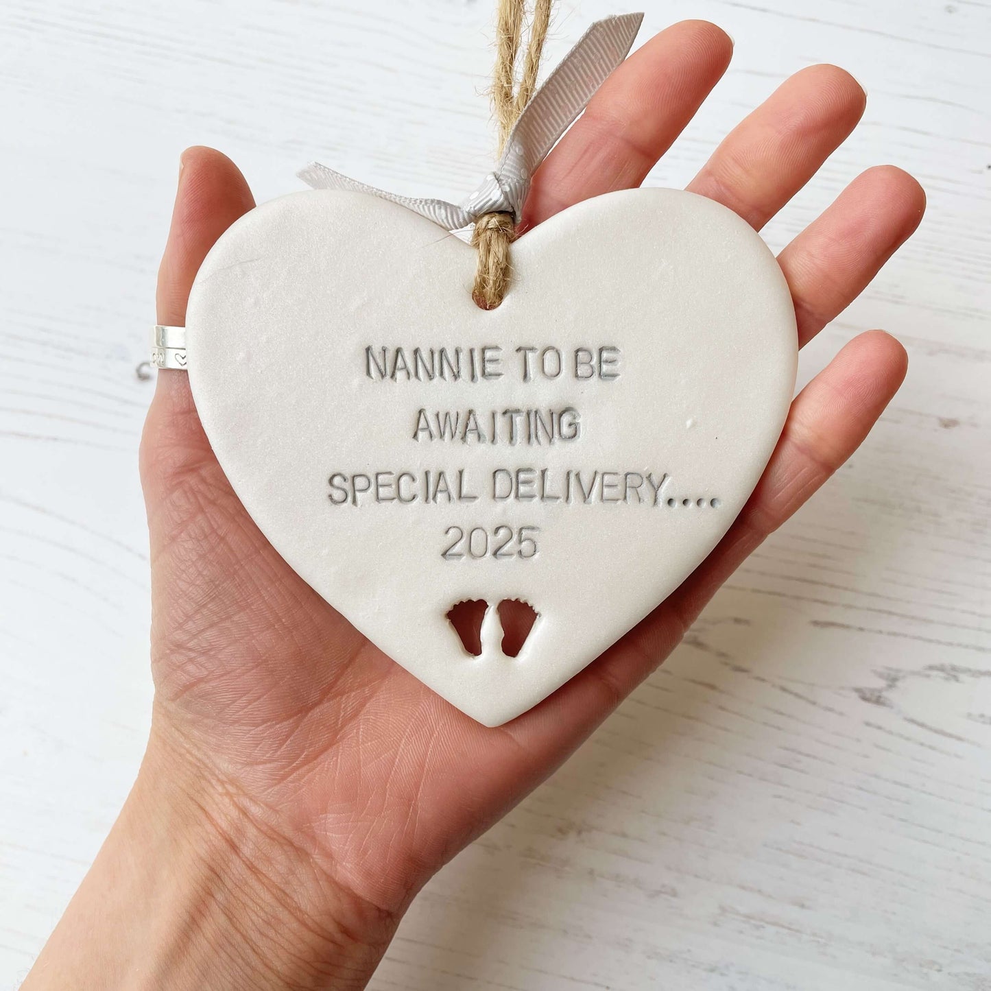 Pearlised white clay hanging heart with baby feet cut out of the bottom and grey personalisation, the heart is personalised with NANNIE TO BE AWAITING SOECIAL DELIVERY…. 2025