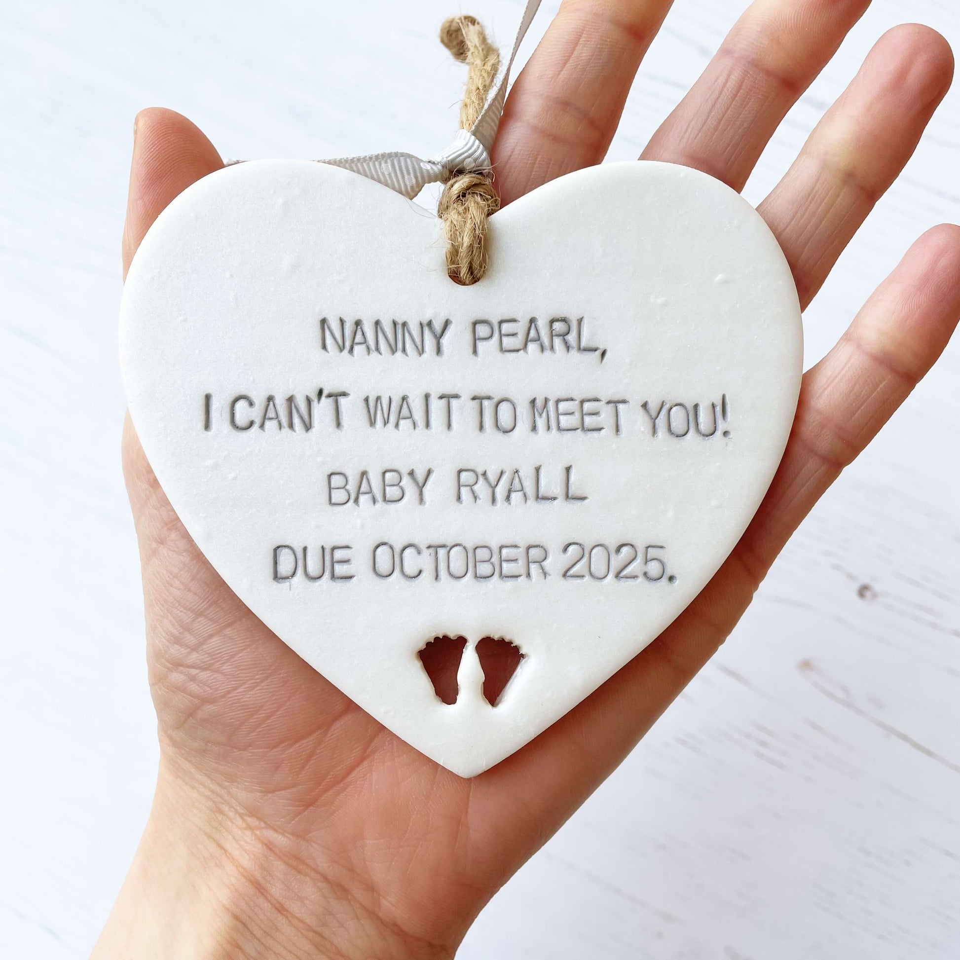 Pearlised white clay hanging heart with baby feet cut out of the bottom and grey personalisation, the heart is personalised with NANNY PEARL, I CAN’T WAIT TO MEET YOU! BABY RYALL DUE OCTOBER 2025