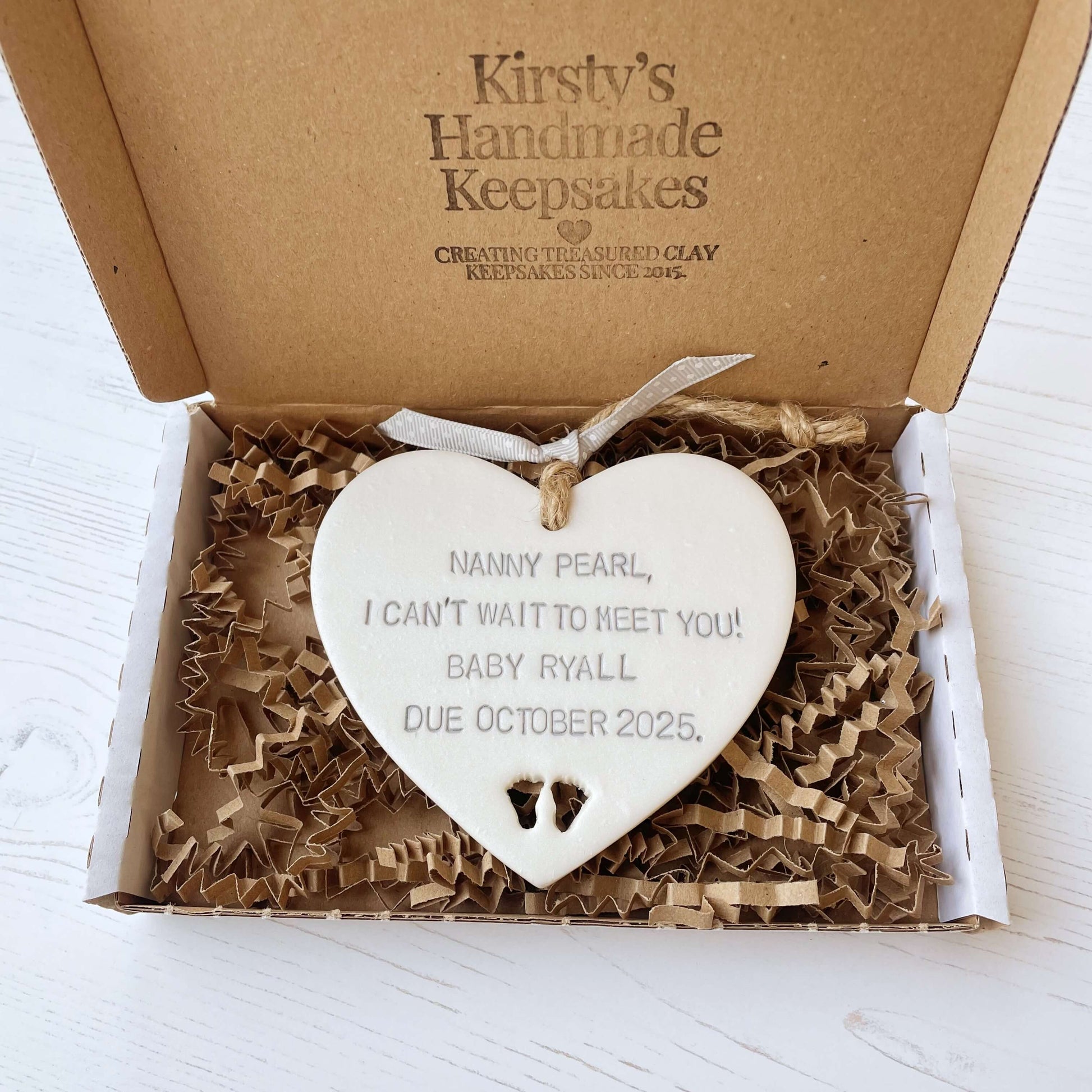 Pearlised white clay hanging heart with baby feet cut out of the bottom and grey personalisation, the heart is personalised with NANNY PEARL, I CAN’T WAIT TO MEET YOU! BABY RYALL DUE OCTOBER 2025