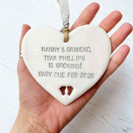 Pearlised white clay hanging heart with baby feet cut out of the bottom and grey personalisation, the heart is personalised with NANNY & GRANDAD TEAM PHILLIPS IS GROWING! BABY DUE FEB 2026