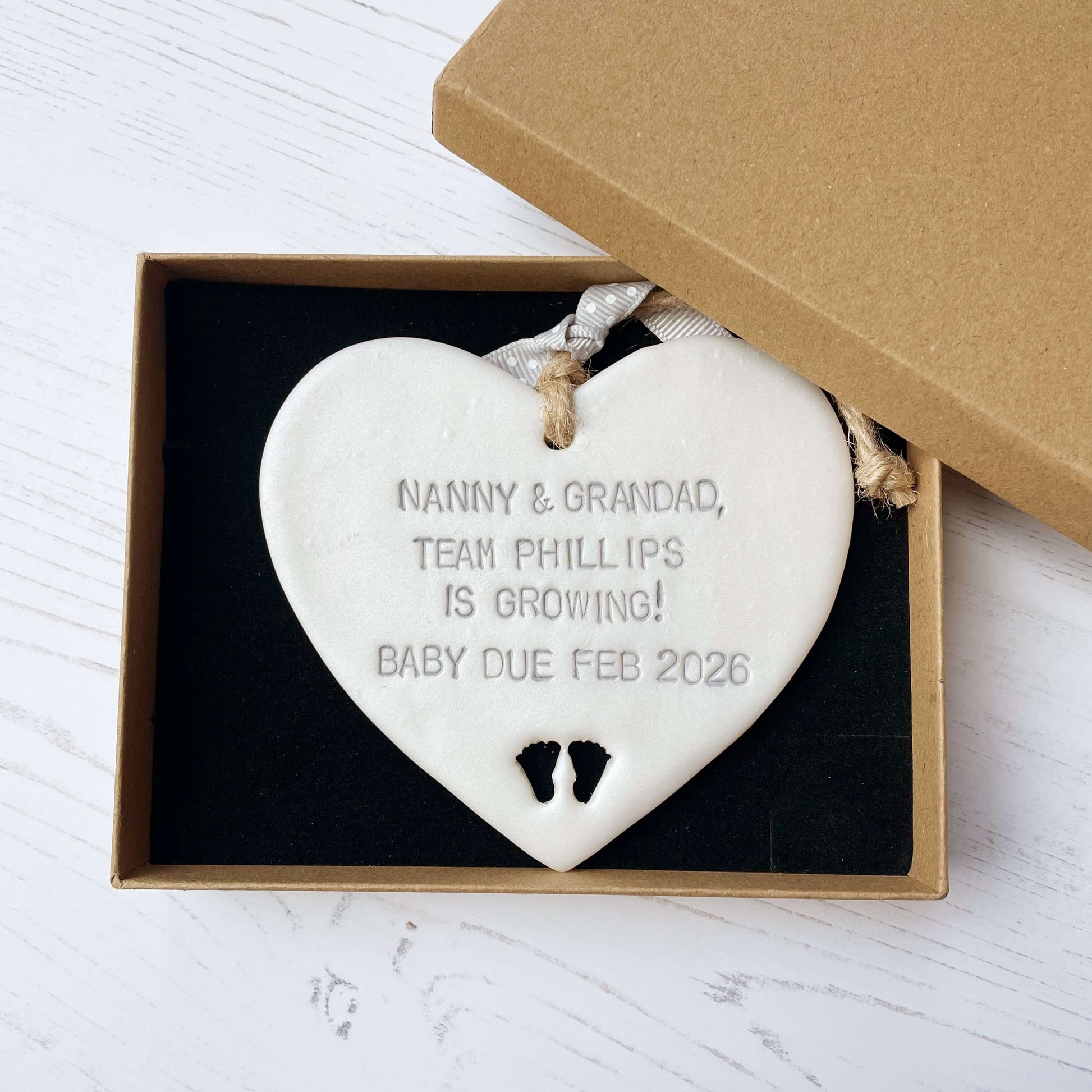 Pearlised white clay hanging heart with baby feet cut out of the bottom and grey personalisation, the heart is personalised with NANNY & GRANDAD TEAM PHILLIPS IS GROWING! BABY DUE FEB 2026