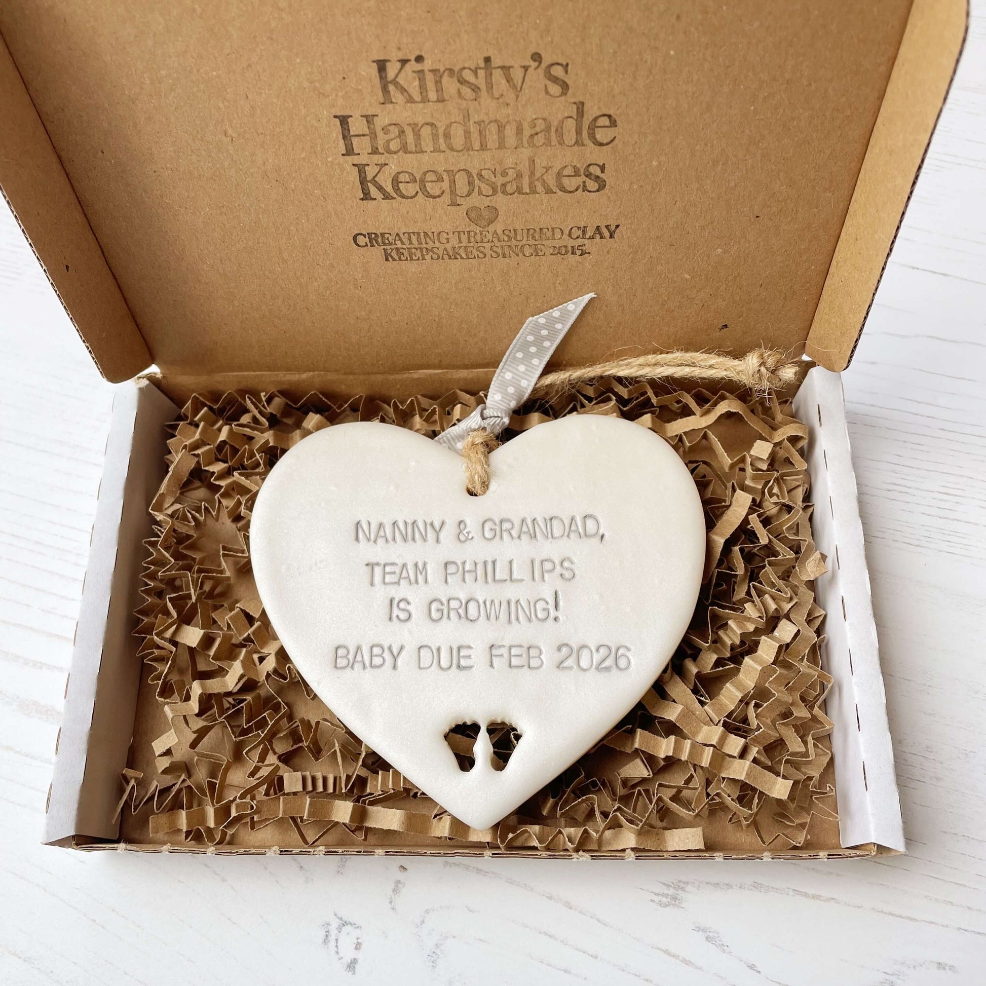 Pearlised white clay hanging heart with baby feet cut out of the bottom and grey personalisation, the heart is personalised with NANNY & GRANDAD TEAM PHILLIPS IS GROWING! BABY DUE FEB 2026