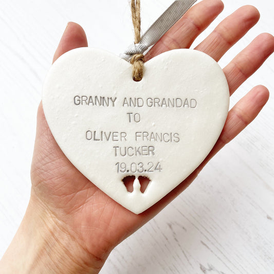 Pearlised white clay hanging heart with baby feet cut out of the bottom and grey personalisation, the heart is personalised with GRANNY AND GRANDAD TO OLIVER FRANCIS TUCKER 19.03.24