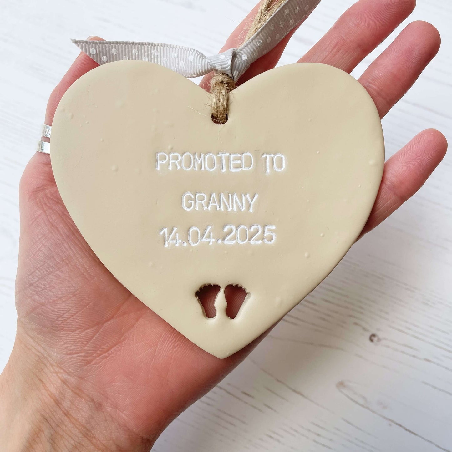 Beige clay hanging heart with baby feet cut out of the bottom and white personalisation, the heart is personalised with PROMOTED TO GRANNY 14.04.2025