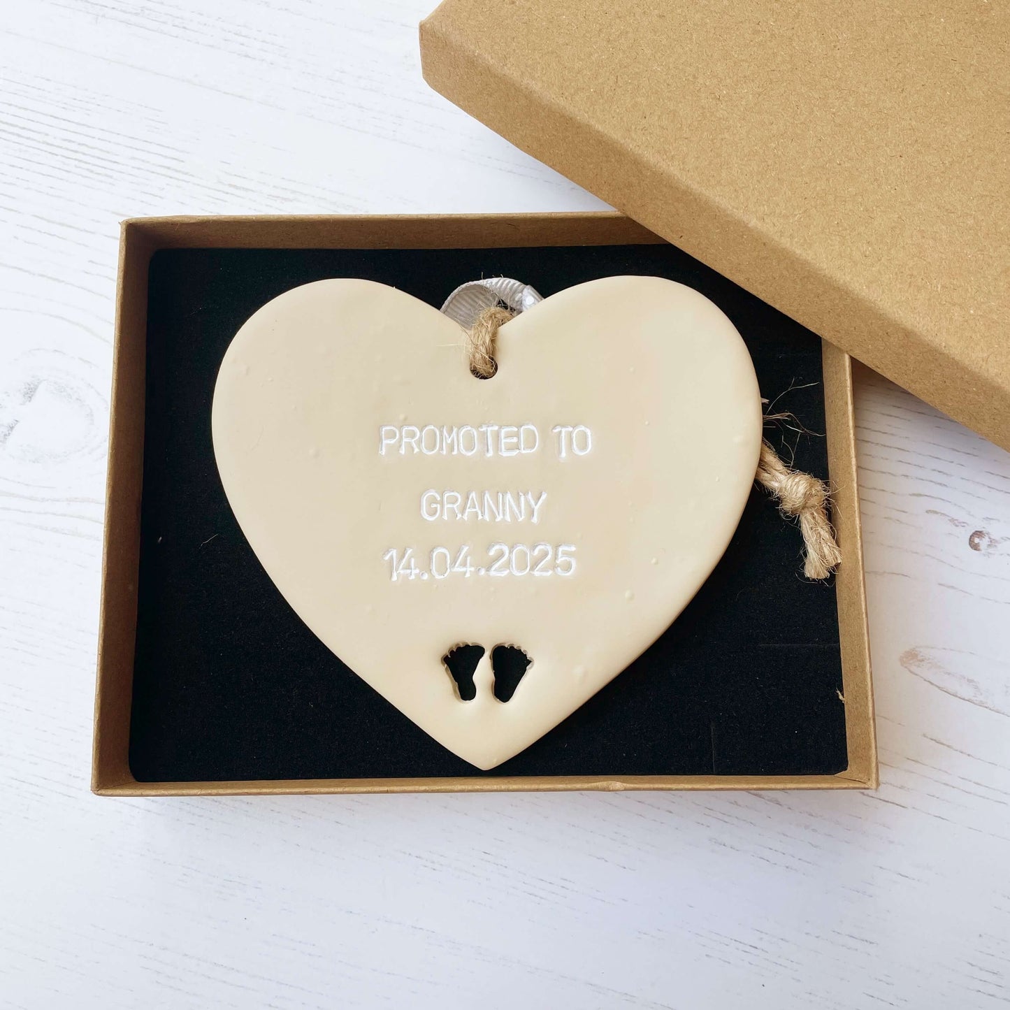 Beige clay hanging heart with baby feet cut out of the bottom and white personalisation, the heart is personalised with PROMOTED TO GRANNY 14.04.2025