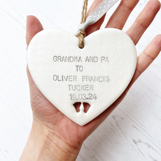 Pearlised white clay hanging heart with baby feet cut out of the bottom and grey personalisation, the heart is personalised with GRANDMA AND PA TO OLIVER FRANCIS TUCKER 19.03.24