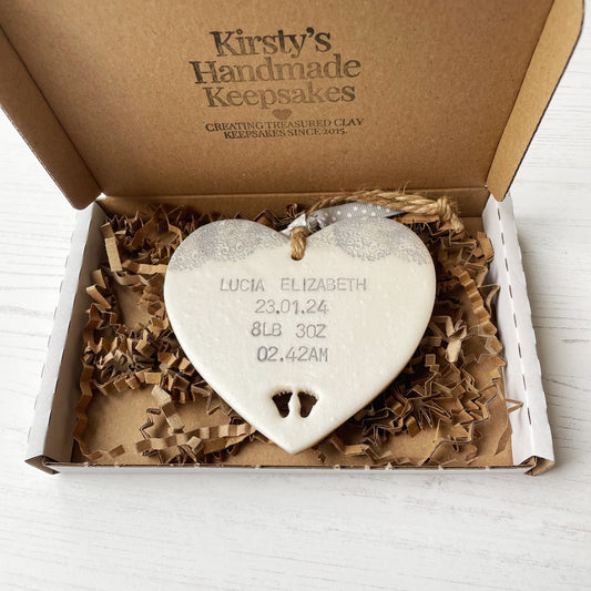 Personalised baby keepsake gift, pearlised white clay hanging heart with baby feet cut out at the bottom of the heart and a grey lace edge at the top of the heart, the heart is personalised with the baby’s name, date of birth, weight and time In a postal box with brown Kraft shredded zigzag paper 