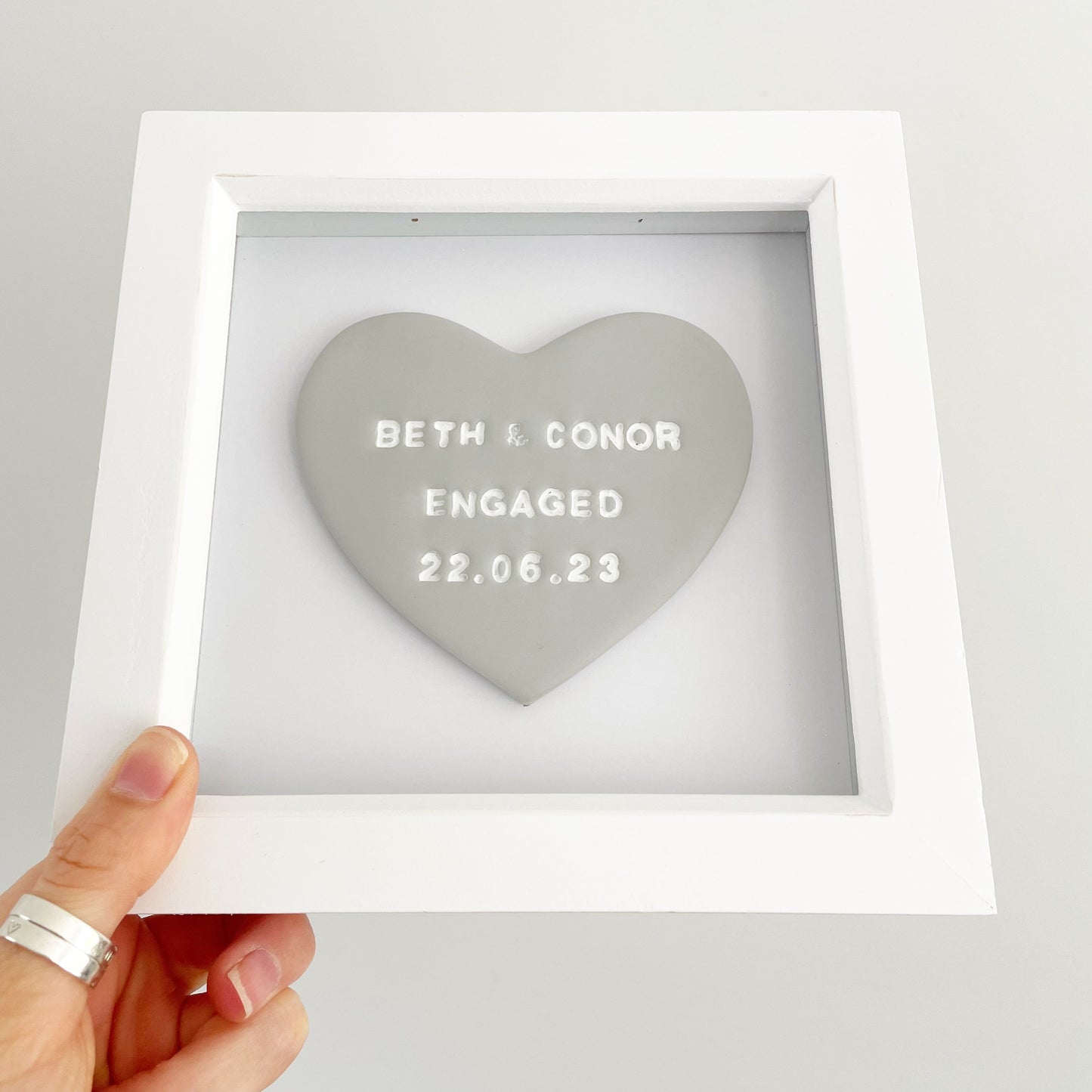 Personalised framed engagement gift, grey clay heart in a white box frame, the heart is personalised with BETH & CONOR ENGAGED 22.06.23