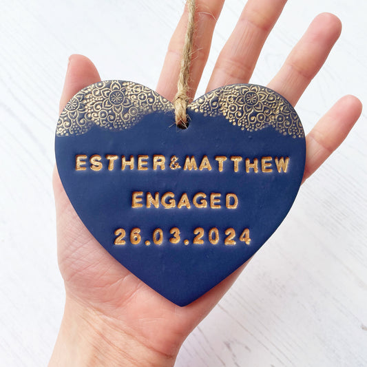 Personalised engagement gift, dark blue clay heart with a gold lace edge at the top of the heart with twine to hang, the heart is personalised with ESTHER & MATTHEW ENGAGED 26.03.2024