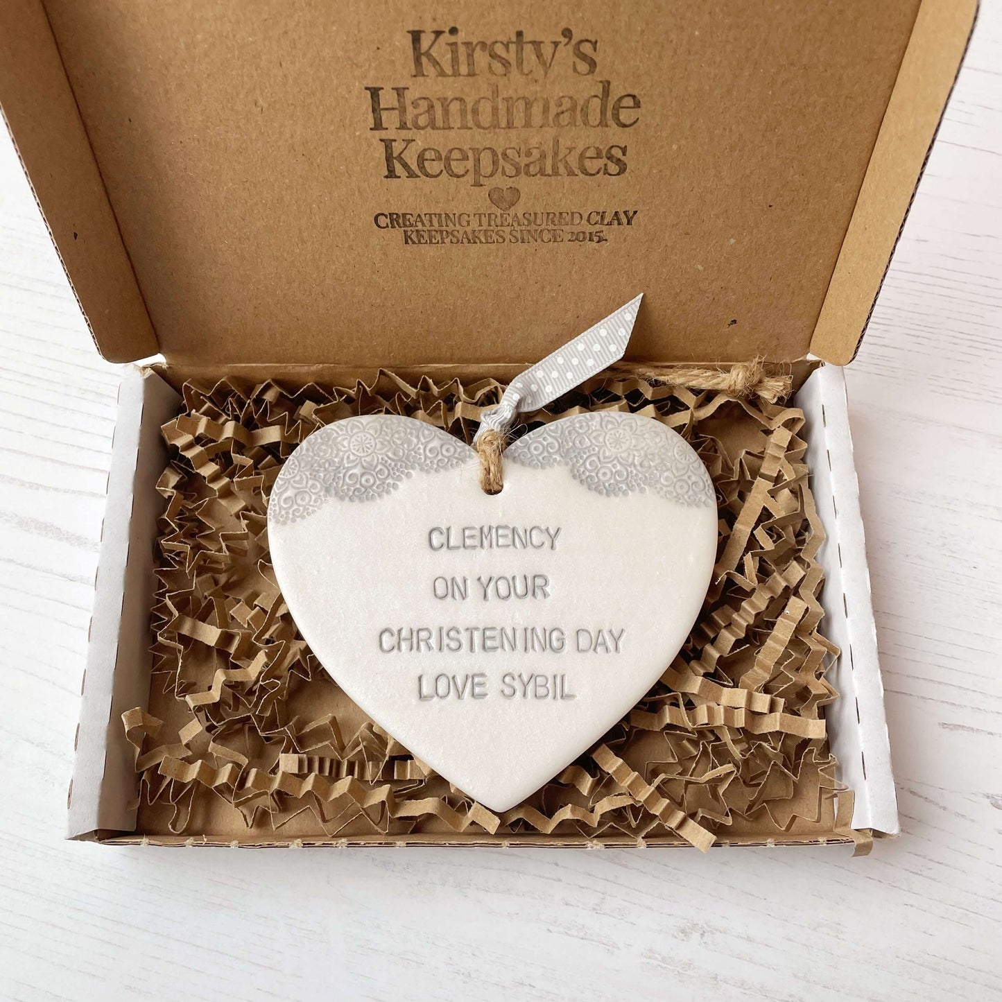 Personalised Christening baptism gift, Pearlised white clay hanging heart with a grey lace edge at the top of the heart, the heart is personalised with CLEMENCY ON YOUR CHRISTENING DAY LOVE SYBIL