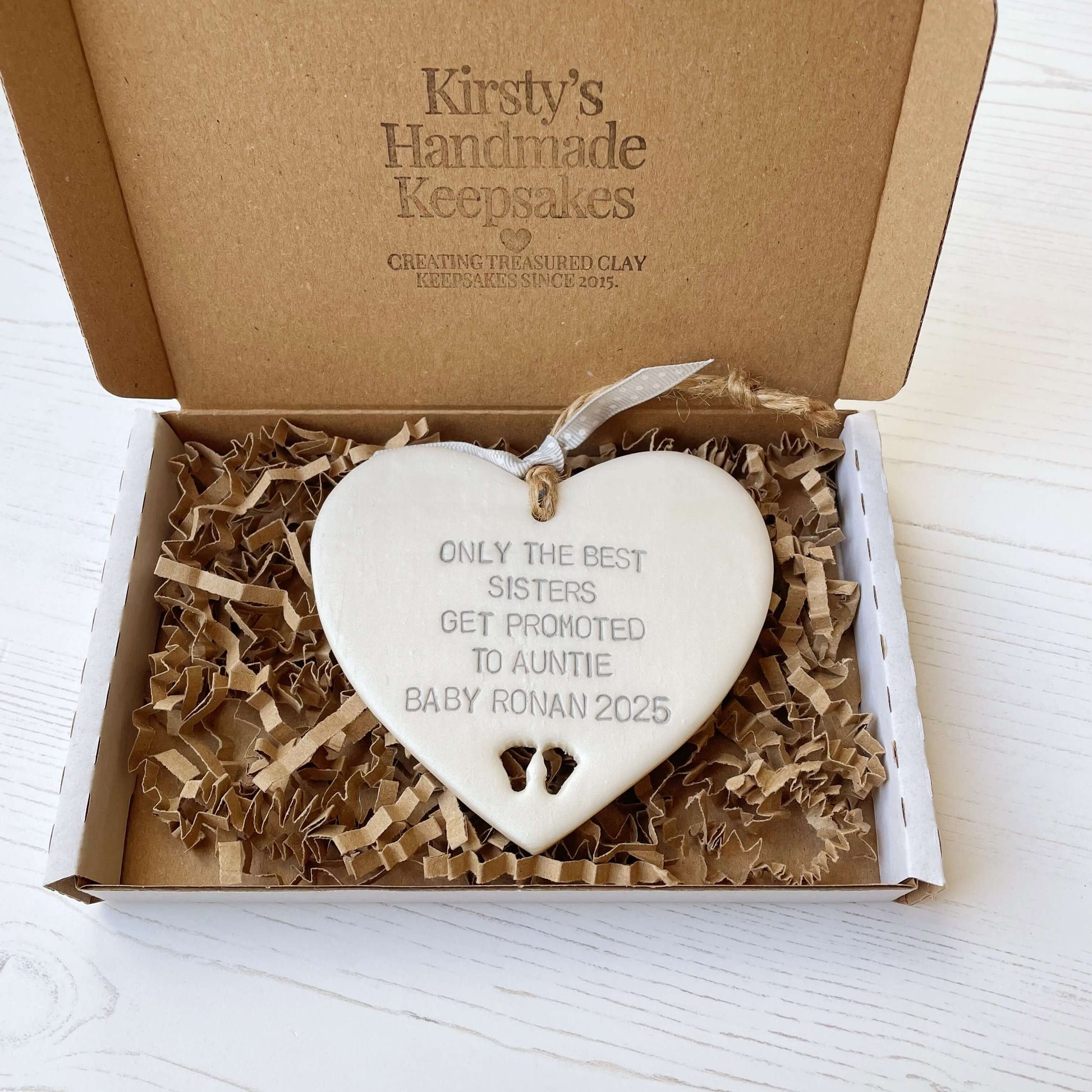 Pearlised white clay hanging heart with baby feet cut out of the bottom and grey personalisation, the heart is personalised with ONLY THE BEST SISTERS GET PROMOTED TO AUNTIE BABY RONAN 2025