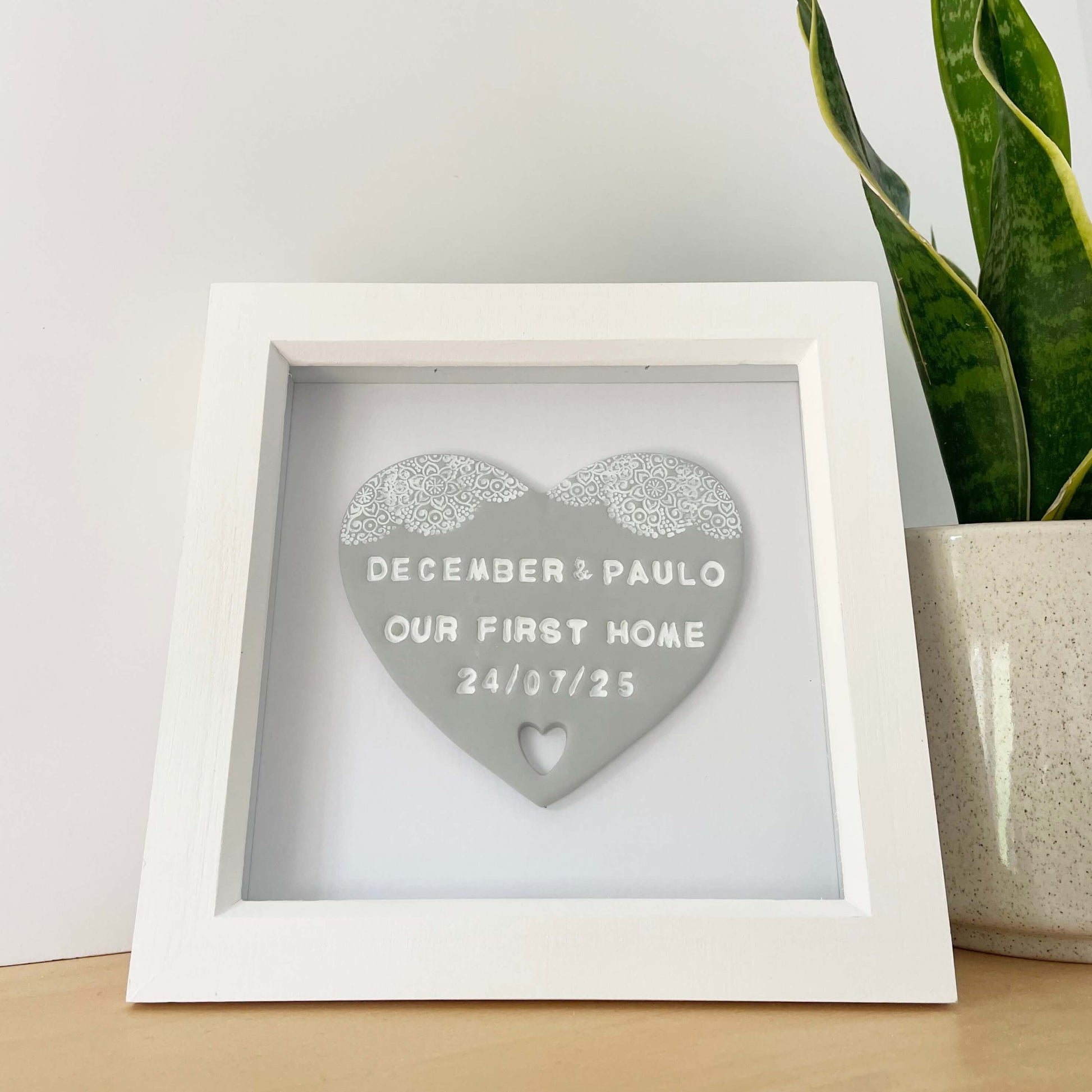 Personalised housewarming new home gift, grey clay heart with a white lace edge at the top of the heart and a heart cut out at the bottom in a white box frame, the heart is personalised with DECEMBER & PAULO OUR FIRST HOME 24/07/25