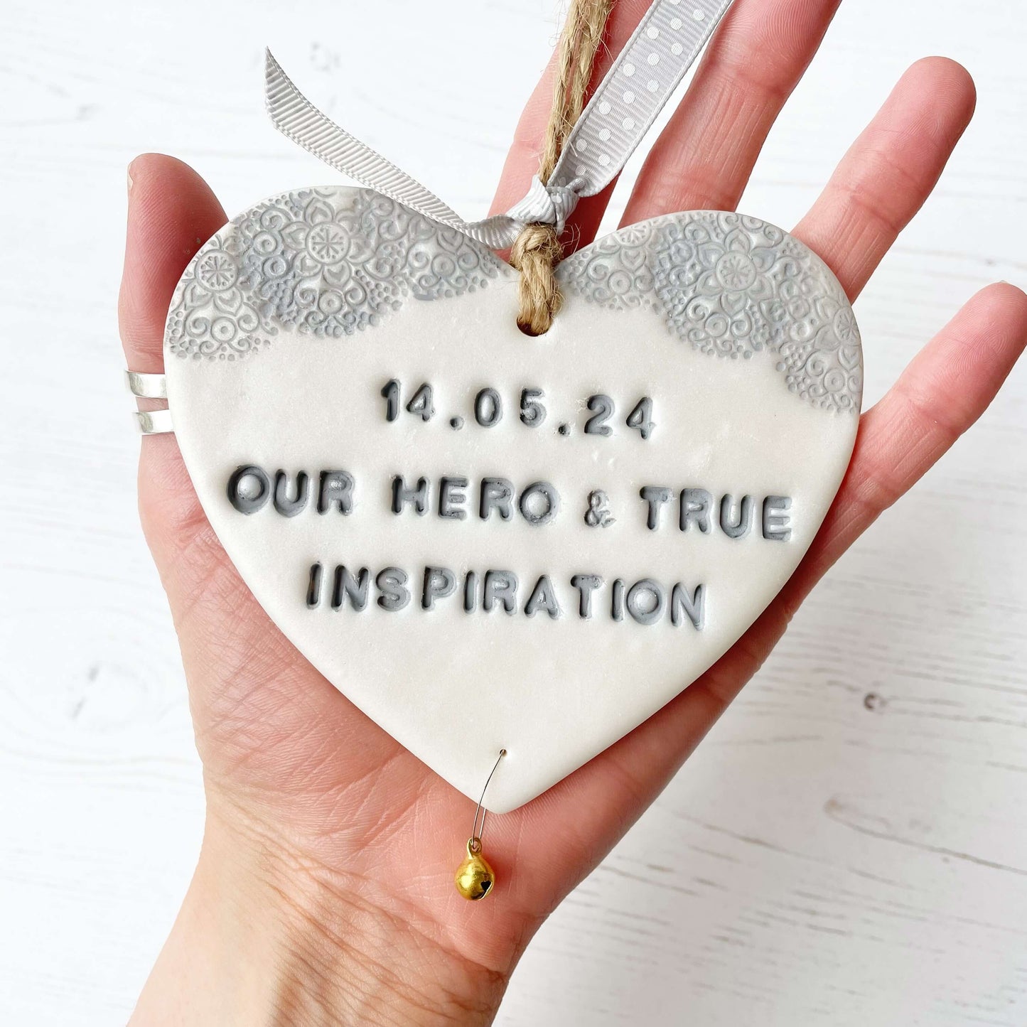 Personalised cancer survivor gift, pearlised white clay hanging heart with a grey lace edge at the top of the heart and a gold bell hanging below, the heart is personalised with 14.05.24 OUR HERO & TRUE INSPIRATION
