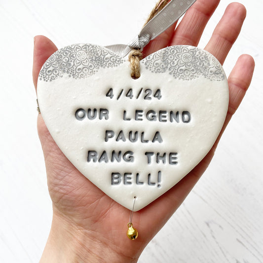 Personalised cancer survivor gift, pearlised white clay hanging heart with a grey lace edge at the top of the heart and a gold bell hanging below, the heart is personalised with 4/4/24 OUR LEGEND PAULA RANG THE BELL!