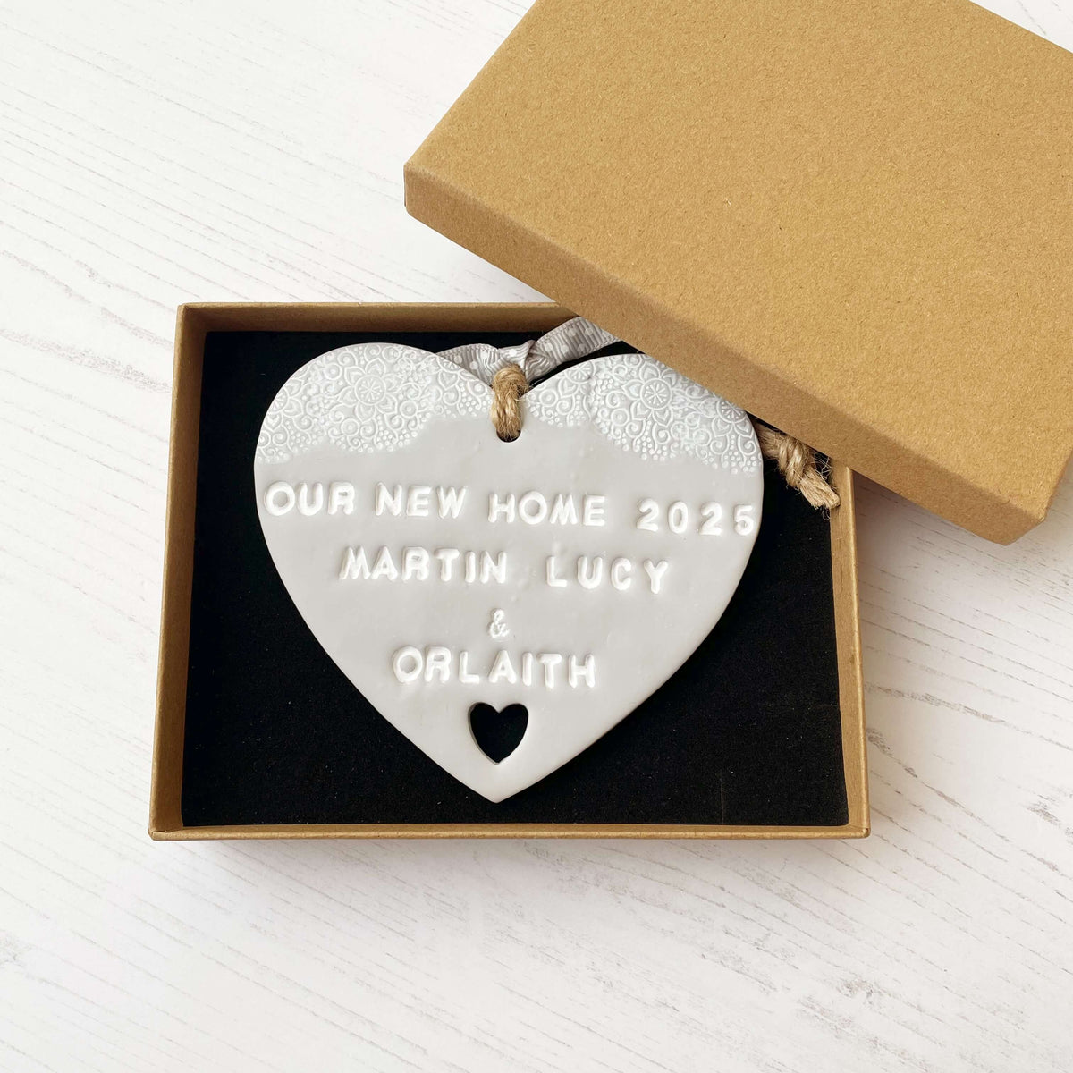 Personalised new home gift grey clay heart with a white lace edge at the top & a heart cut out at the bottom with twine for hanging personalised with OUR NEW HOME 2025 MARTIN LUCY & ORLAITH