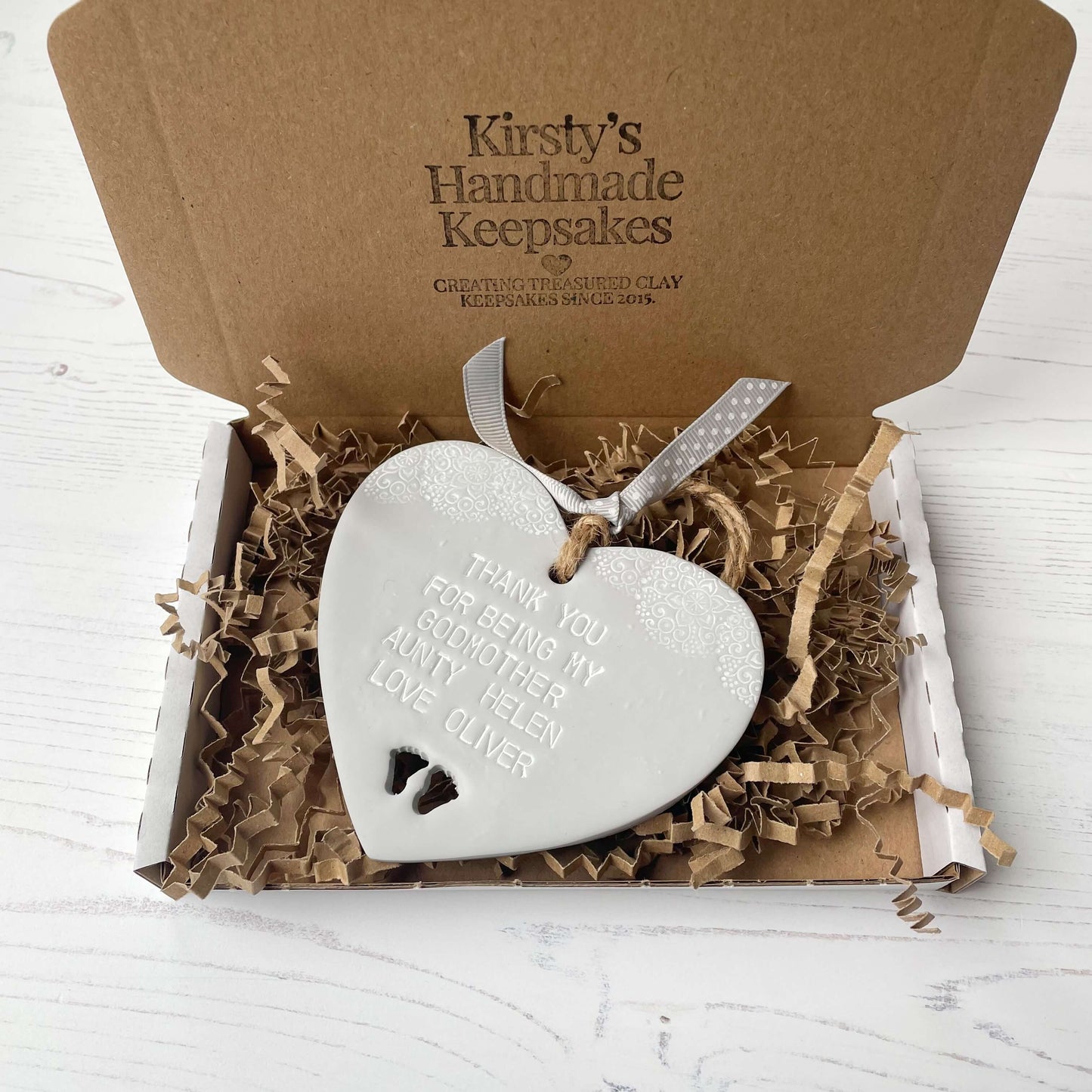 Personalised Godmother gift, grey clay hanging heart with baby feet cut out of the bottom and white lace edge at the top of the heart, the heart is personalised with THANK YOU FOR BEING MY GODMOTHER HELEN LOVE OLIVER