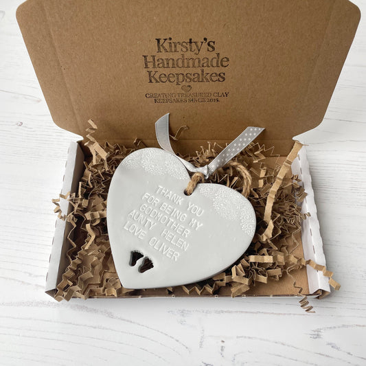 Personalised Godmother gift, grey clay hanging heart with baby feet cut out of the bottom and white lace edge at the top of the heart, the heart is personalised with THANK YOU FOR BEING MY GODMOTHER HELEN LOVE OLIVER. In a white postal box with brown Kraft zigzag shredded paper