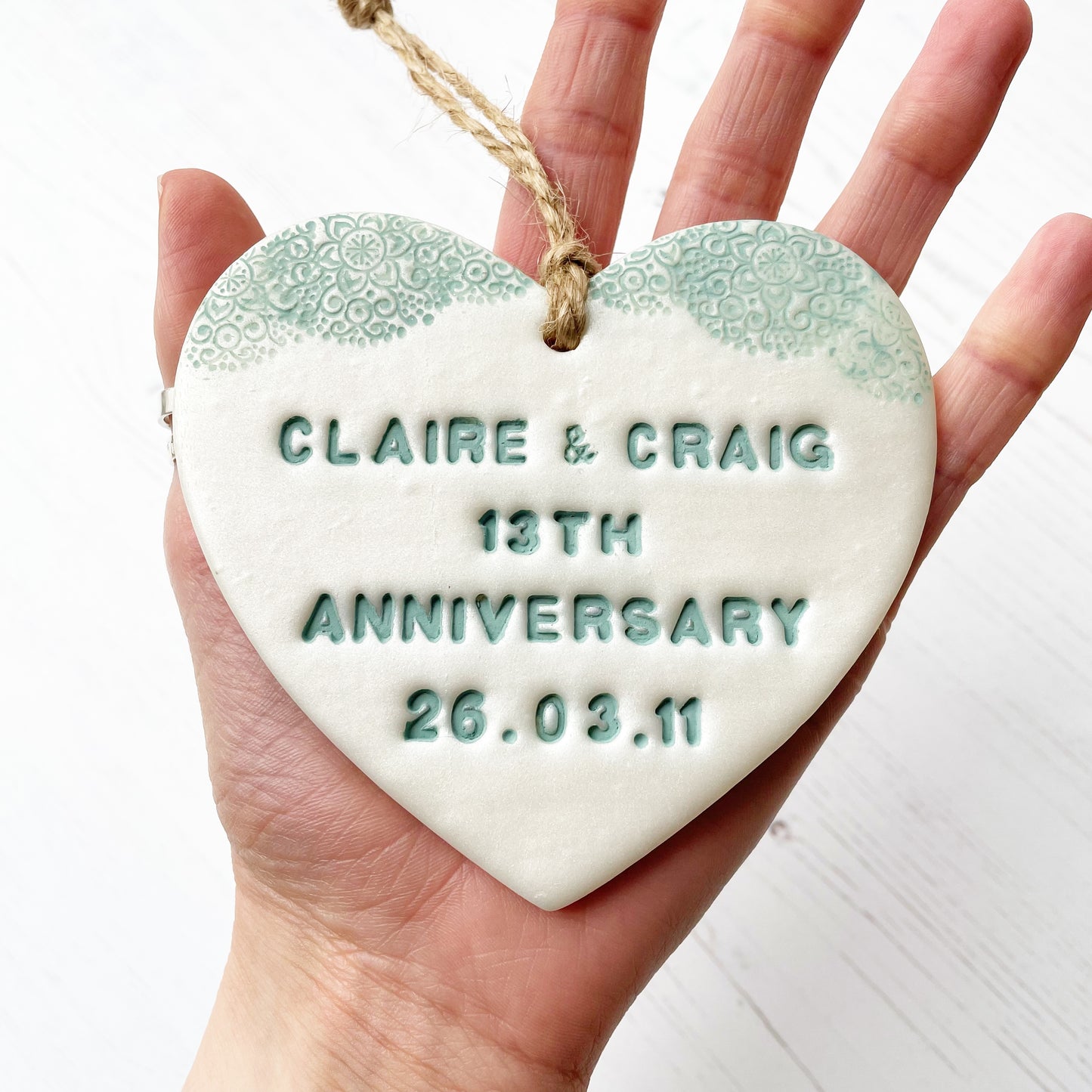 Personalised 13th wedding anniversary gift, pearlised white clay hanging heart with a sage green lace edge at the top of the heart, the heart is personalised with CLAIRE & CRAIG 13TH ANNIVERSARY 26.03.11