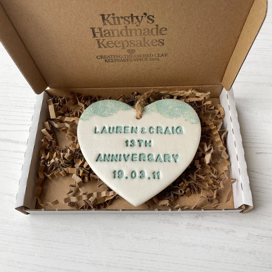 Personalised 13th wedding anniversary gift, pearlised white clay hanging heart with a sage green lace edge at the top of the heart, the heart is personalised with LAUREN & CRAIG 13TH ANNIVERSARY 19.03.11 In a white postal box with shredded zigzag Kraft paper