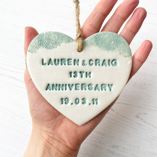 Personalised 13th wedding anniversary gift, pearlised white clay hanging heart with a sage green lace edge at the top of the heart, the heart is personalised with LAUREN & CRAIG 13TH ANNIVERSARY 19.03.11