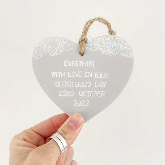 Personalised Christening baptism gift, grey clay hanging heart with a white lace edge at the top of the heart, the heart is personalised with EVRITHIKI WITH LOVE ON YOUR CHRISTENING DAY 22ND OCTOBER 2022