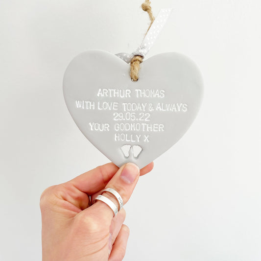 Personalised Christening gift, grey clay hanging heart with baby feet cut out of the bottom, the heart is personalised with  ARTHUR THOMAS WITH LOVE TODAY & ALWAYS 29.05.22 YOUR GODMOTHER HOLLY X