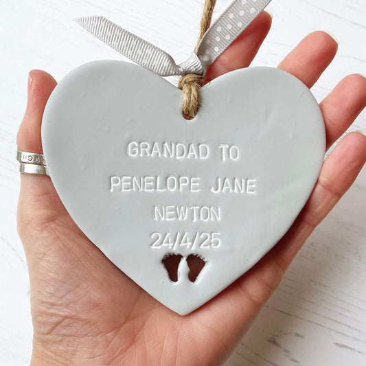 Grey clay hanging heart with baby feet cut out of the bottom and white personalisation, the heart is personalised with GRANDAD TO PENELOPE JANE NEWTON 24/4/25