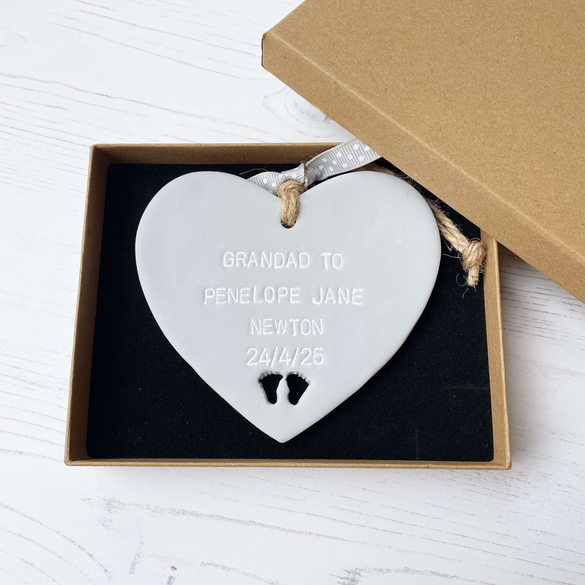 Grey clay hanging heart with baby feet cut out of the bottom and white personalisation, the heart is personalised with GRANDAD TO PENELOPE JANE NEWTON 24/4/25