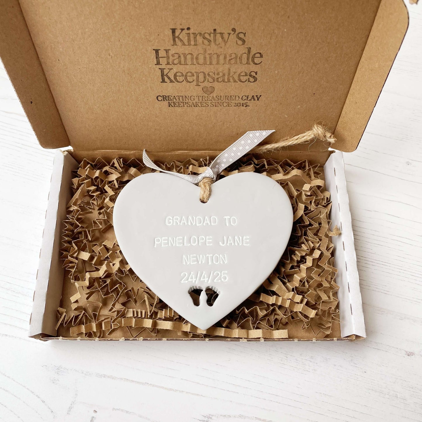 Grey clay hanging heart with baby feet cut out of the bottom and white personalisation, the heart is personalised with GRANDAD TO PENELOPE JANE NEWTON 24/4/25