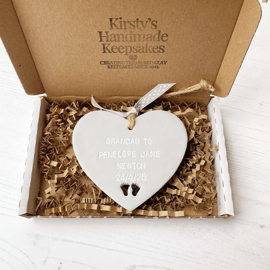 Grey clay hanging heart with baby feet cut out of the bottom and white personalisation, the heart is personalised with GRANDAD TO PENELOPE JANE NEWTON 24/4/25