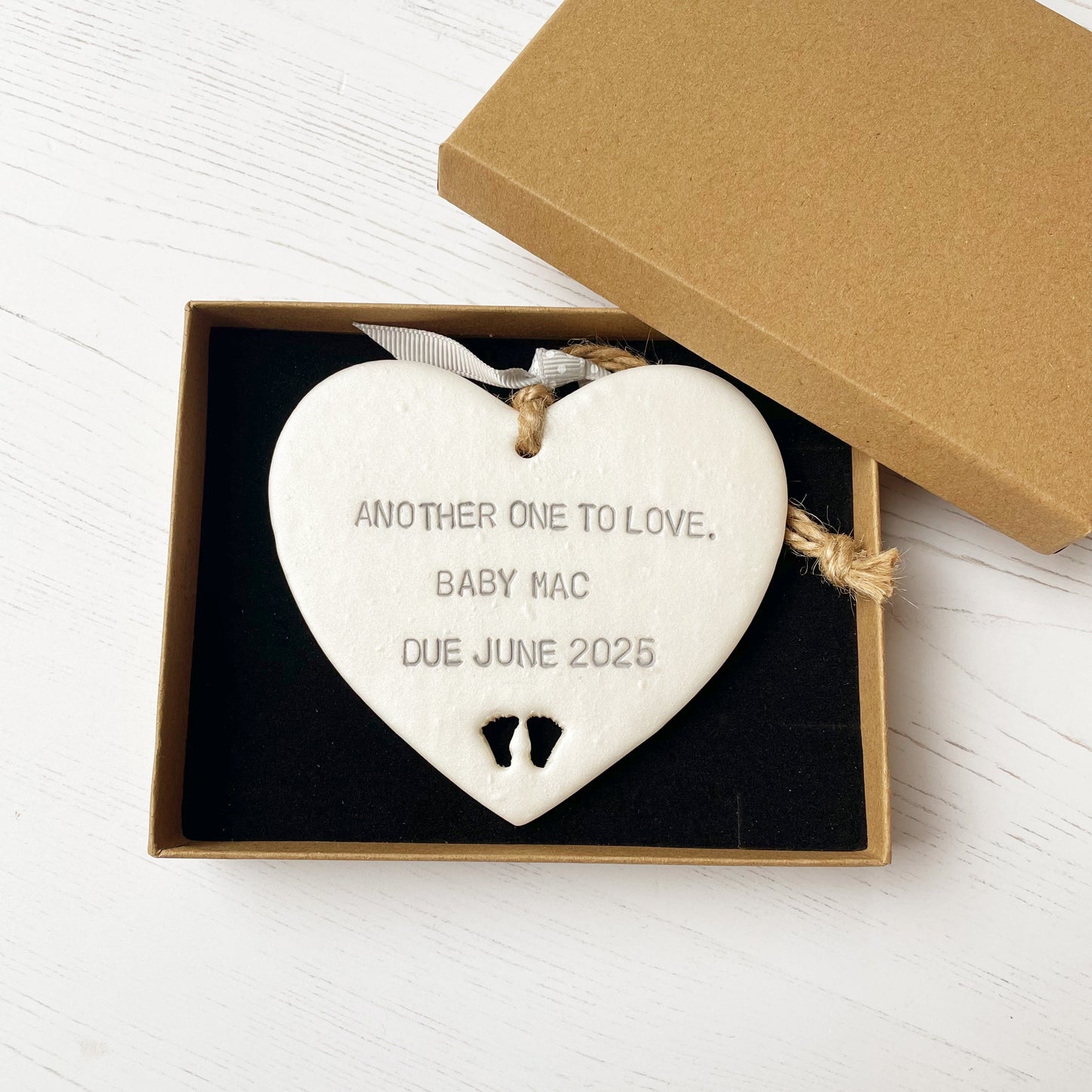 Pearlised white clay hanging heart with baby feet cut out of the bottom and grey personalisation, the heart is personalised with ANOTHER ONE TO LOVE. BABY MAC DUE JUNE 2025