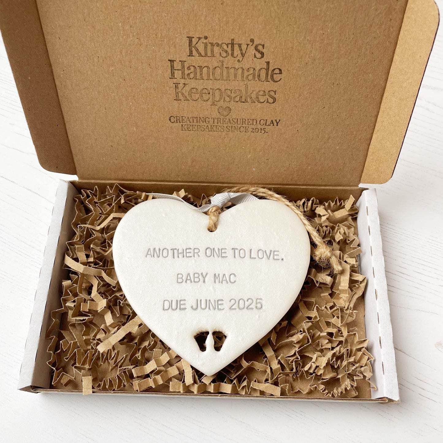 Pearlised white clay hanging heart with baby feet cut out of the bottom and grey personalisation, the heart is personalised with ANOTHER ONE TO LOVE. BABY MAC DUE JUNE 2025