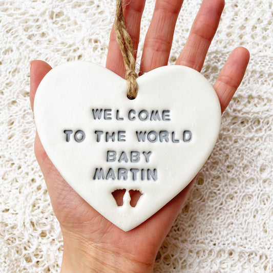 Personalised baby announcement heart sign, in pearlised white clay hanging heart with a grey writing and baby feet cut out at the bottom of the heart, the heart is personalised with WELCOME TO THE WORLD BABY MARTIN