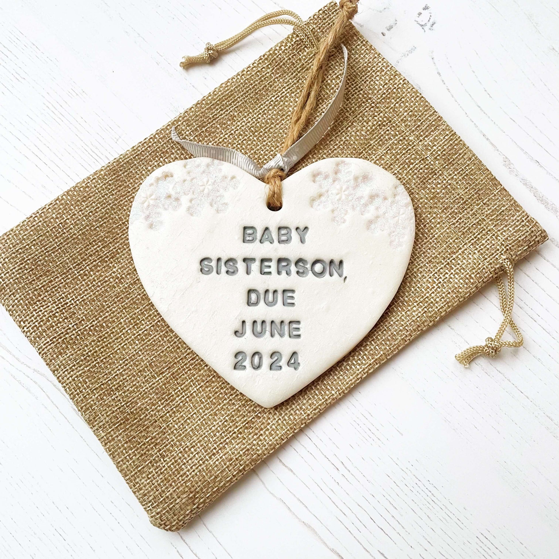 Personalised baby's first Christmas heart ornament, pearlised white clay with BABY SISTERSON, DUE JUNE 2024 painted grey, decorated with 2 iridescent glitter snowflakes on either side of the top of the heart