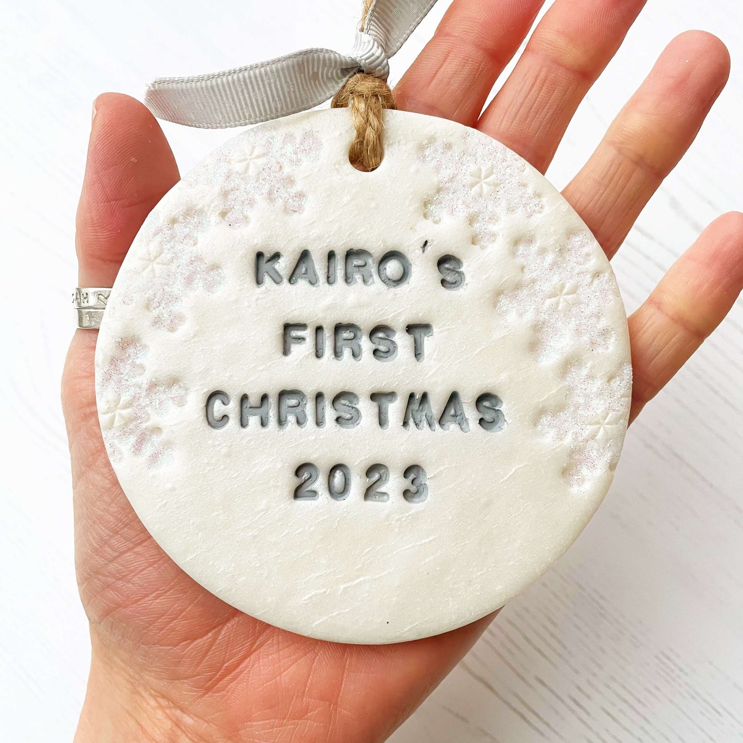 Personalised baby’s first Christmas bauble ornament, pearlised white clay round personalised with Kairo’s first Christmas 2023 in grey paint, decorated with 3 iridescent glitter snowflakes on either side of the bauble